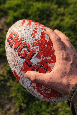 Hand holding a white and red Optimum England rugby ball with ENGLAND print above grass - Optimum