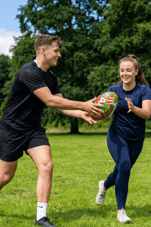 Two smiling players passing a red, green and white Optimum Wales rugby ball in a park - Optimum