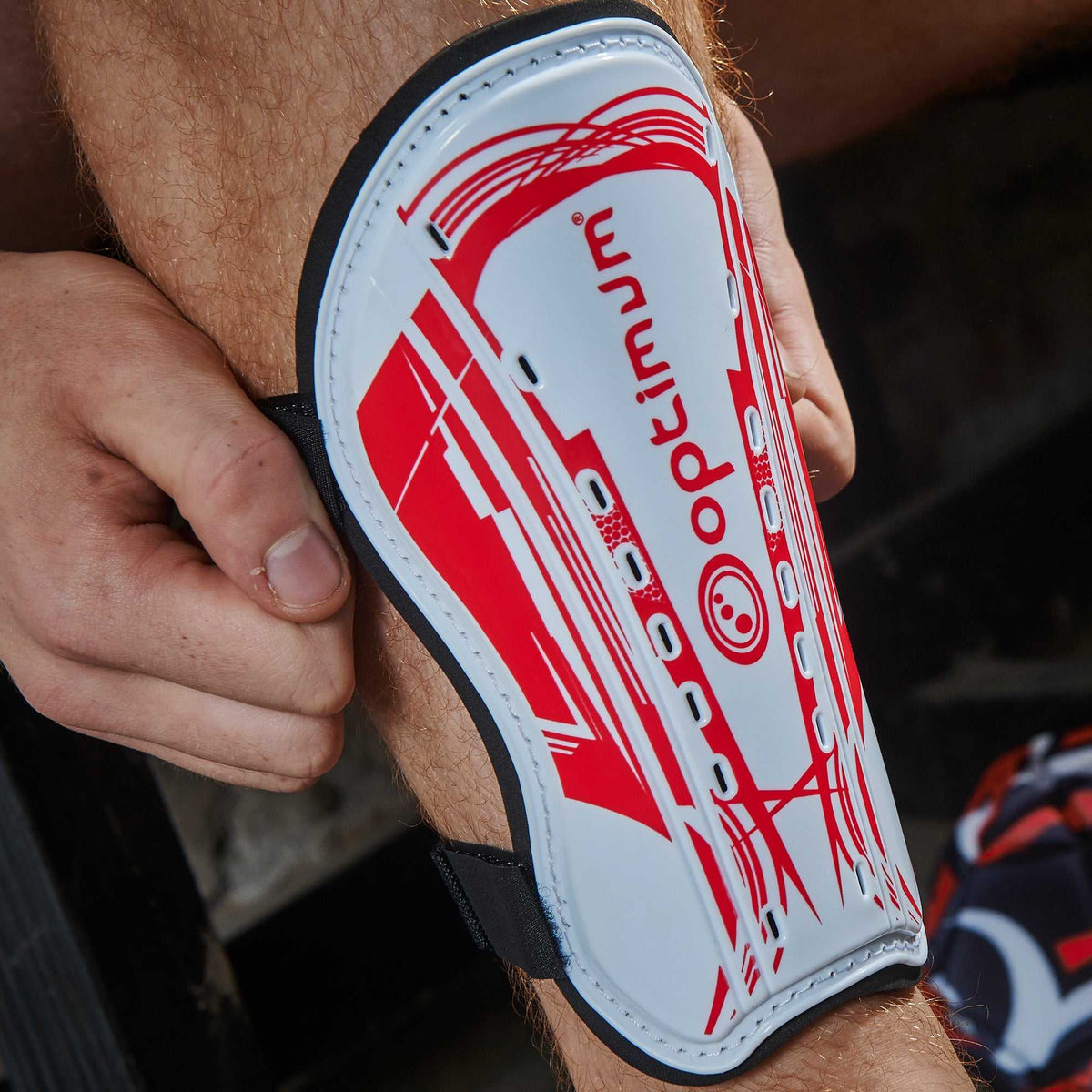 Hand holding white and red Optimum Velocidade shin guard with red print and logo against leg - Optimum